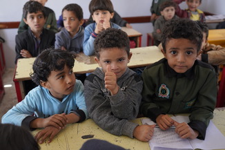 Boys sitting three to a desk in Yemen. One giving a thumbs up to the camera.