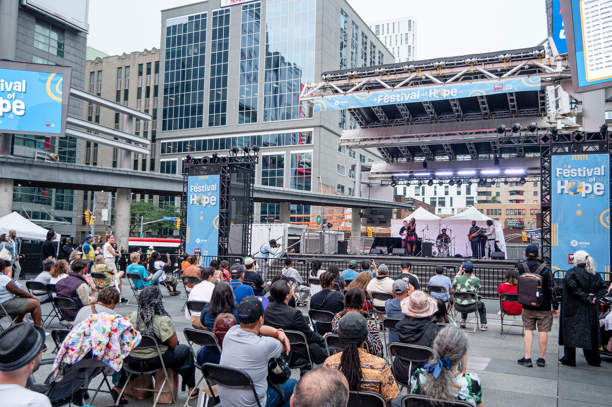 A crowd gathered at a large stage in Yonge-Dundas Square, Toronto. Digital billboards and posters have A Festival of Hope banners in blue and yellow. SingerAmanie Illfated performs on stage with her band.