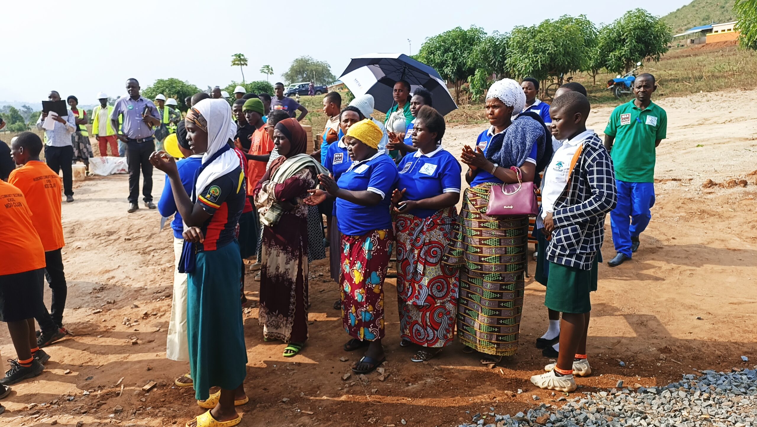 Community members attend a grand opening of a new accelerated learning program. Standing in a group. Some women in blue shirts support young mothers attending school by caring for their children.
